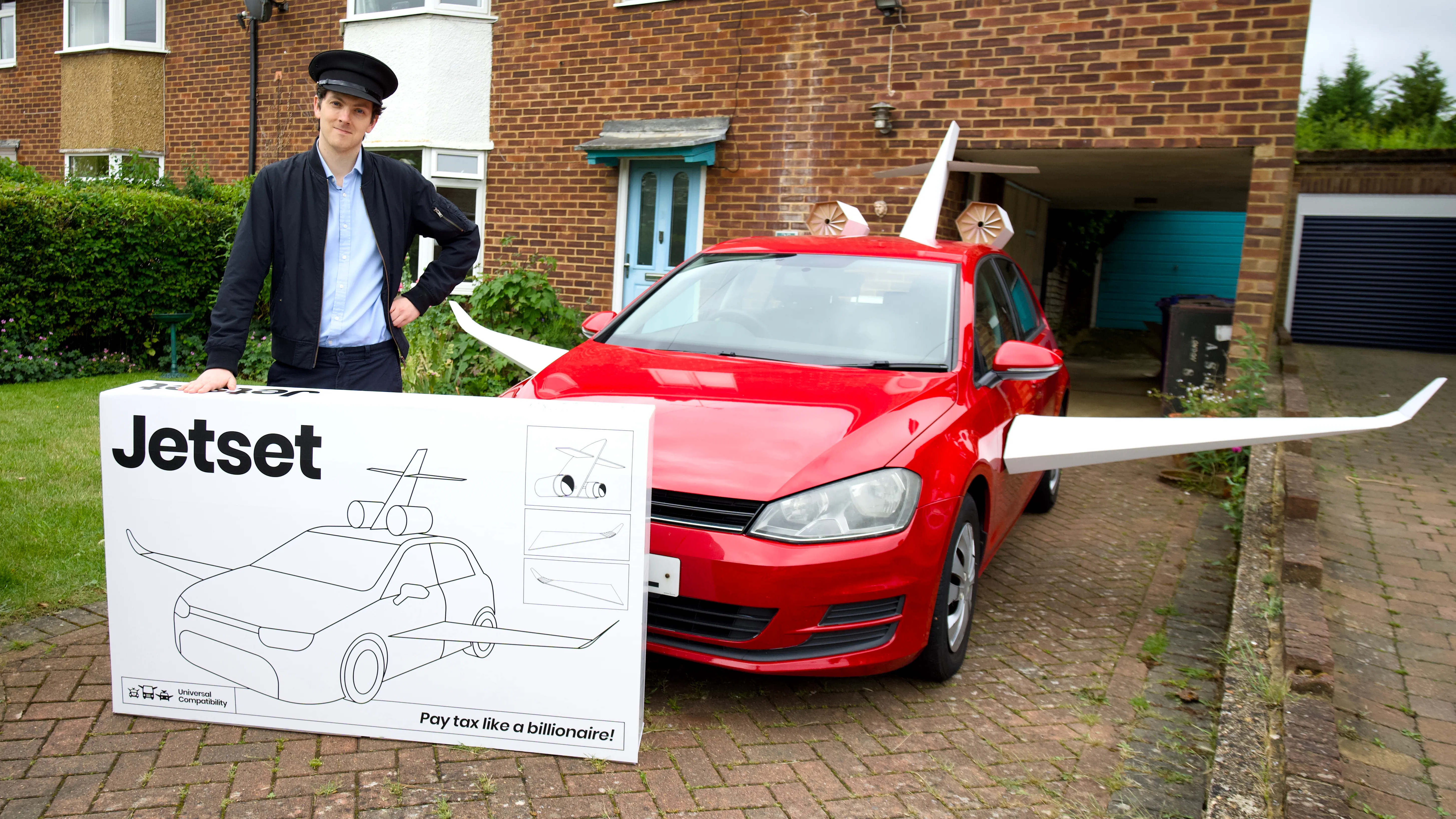 A red hatchback fitted with cardboard wings, beside a chauffeur and a flat-pack Jetset box